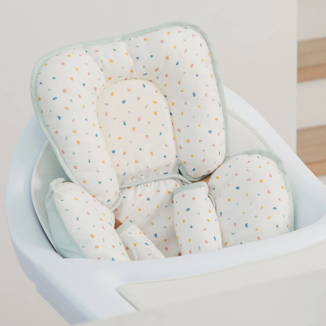 ClevaMama ClevaFoam Head and Neck Support Terrazzo design — baby head and body cushion fitted in a high chair. Ergonomic newborn support pillow made from ClevaFoam for superior comfort and flat head syndrome prevention. Breathable, soft, and colorful muslin cover ideal for car seats, bouncers, strollers, and feeding chairs