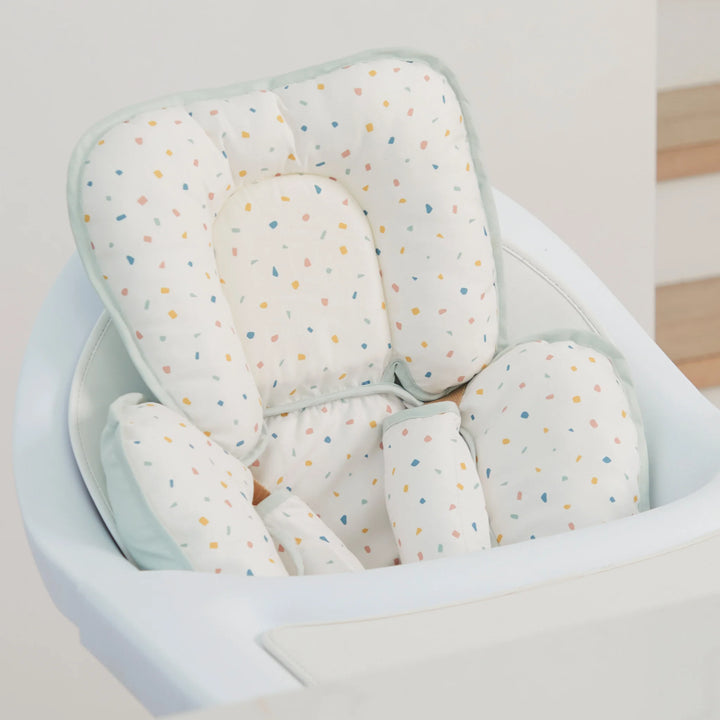 ClevaMama ClevaFoam Head and Neck Support Terrazzo design — baby head and body cushion fitted in a high chair. Ergonomic newborn support pillow made from ClevaFoam for superior comfort and flat head syndrome prevention. Breathable, soft, and colorful muslin cover ideal for car seats, bouncers, strollers, and feeding chairs