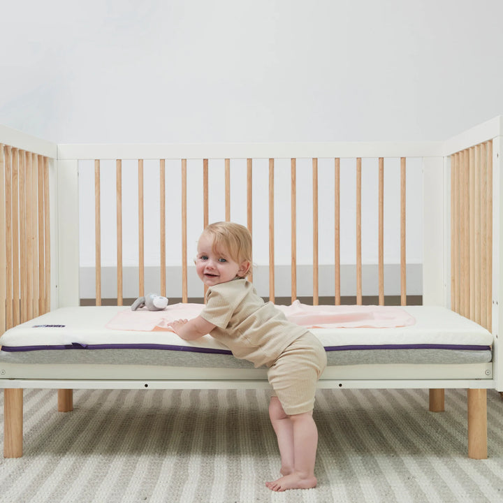 Smiling toddler standing in cot on Clevamama ClevaFoam Waterproof Support Mattress – breathable, hypoallergenic, and durable baby mattress providing ultimate comfort and spinal support, featuring waterproof protection and removable washable cover for a safe, hygienic sleep environment.