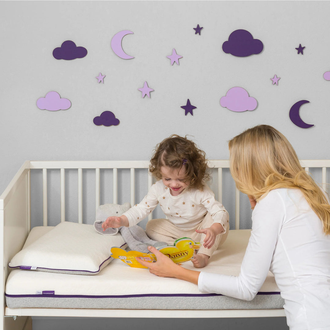 Mother reading to toddler sitting on Clevamama Waterproof Support Mattress in cot bed – premium breathable baby mattress with waterproof protection, hypoallergenic reflex support foam, and removable washable cover for safe, comfortable sleep and play.