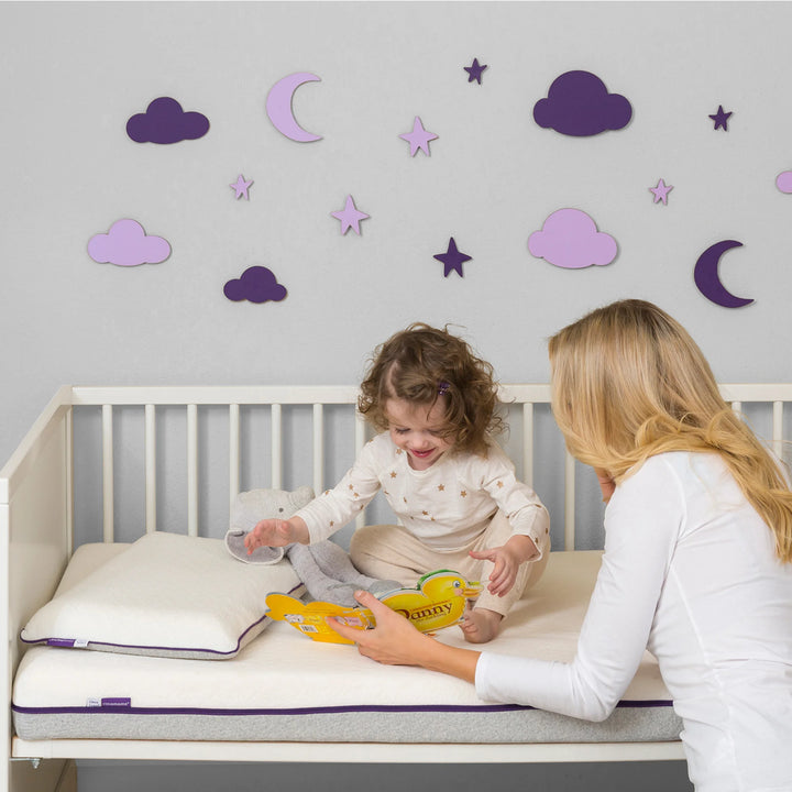 Mother reading to toddler sitting on Clevamama Waterproof Support Mattress in cot bed – premium breathable baby mattress with waterproof protection, hypoallergenic reflex support foam, and removable washable cover for safe, comfortable sleep and play.
