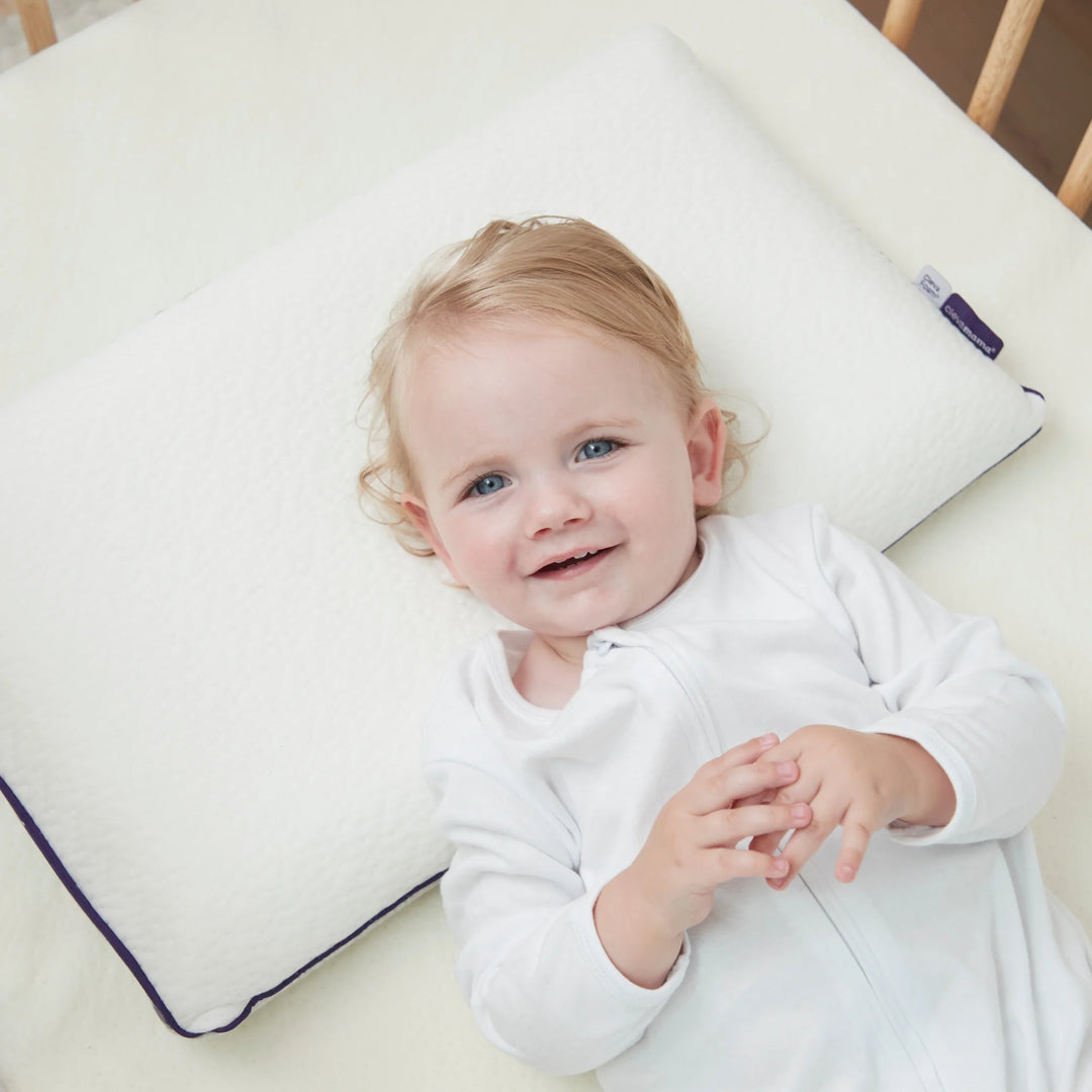 Smiling toddler resting on Clevamama ClevaFoam® Toddler Pillow – breathable, hypoallergenic memory foam pillow designed for children aged 12 months and up. Provides optimal neck and head support for safe, comfortable sleep and healthy spine alignment in a cot or toddler bed.