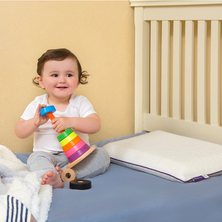 Smiling toddler playing beside Clevamama ClevaFoam® Toddler Pillow on bed – breathable, hypoallergenic memory foam pillow for children aged 12 months and up. Designed to support healthy spine alignment, prevent flat head syndrome, and provide soft, safe comfort for naps and nighttime sleep.