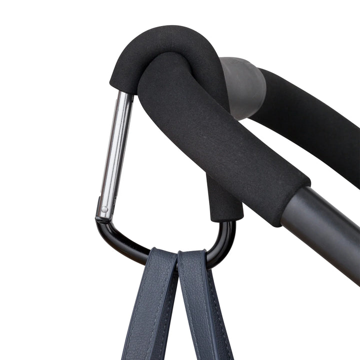 ClevaMama ClevaHooks stroller clip attached to a stroller handle, holding a bag strap with the carabiner hook.