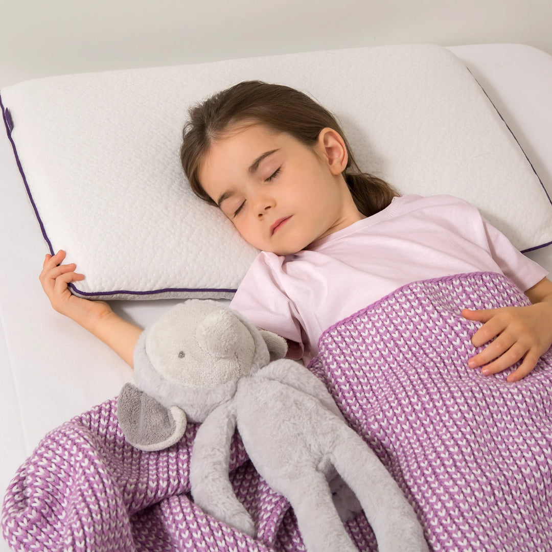 A young child sleeping peacefully on a white ClevaMama Junior Pillow, covered with a purple knit blanket and holding a soft grey stuffed toy.