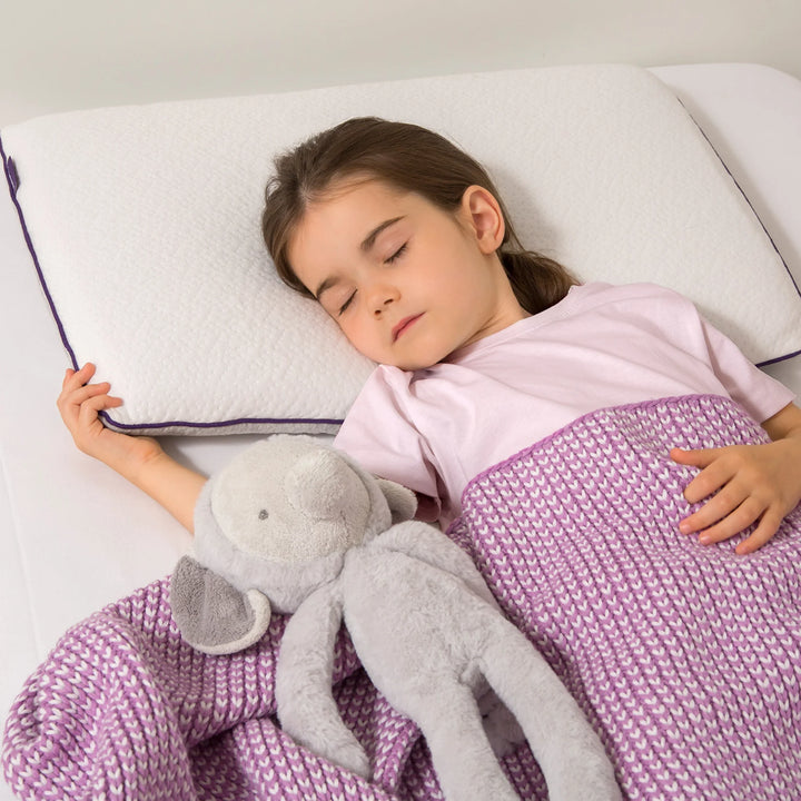 A young child sleeping peacefully on a white ClevaMama Junior Pillow, covered with a purple knit blanket and holding a soft grey stuffed toy.