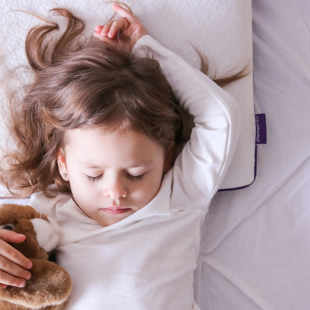 A young child sleeping on a white ClevaMama Junior Pillow with their arms raised above their head, holding a soft brown teddy bear.