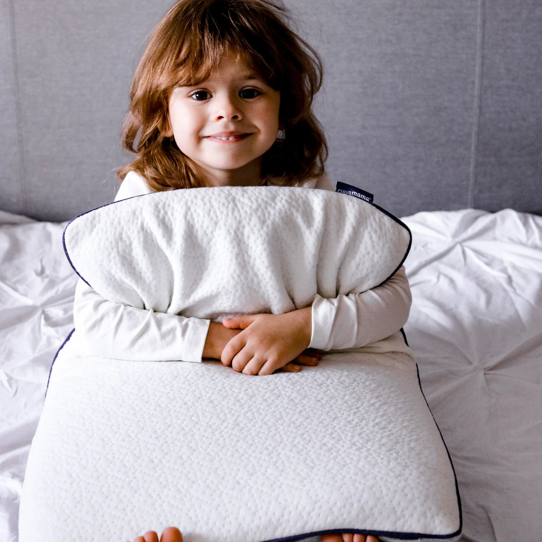 A young child sitting on a bed, smiling while hugging a white ClevaMama Junior Pillow in front of them.