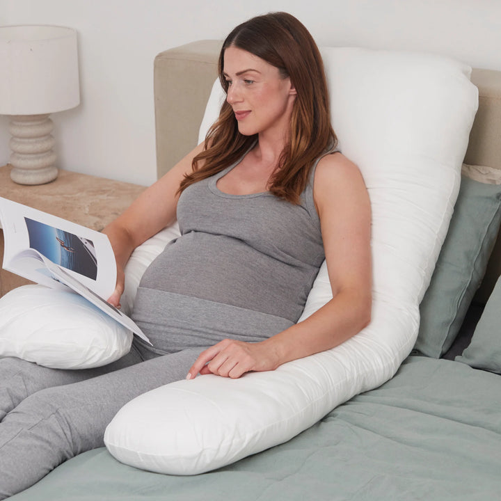 “Pregnant woman sitting up in bed reading a magazine while supported by a white U-shaped maternity pillow.”