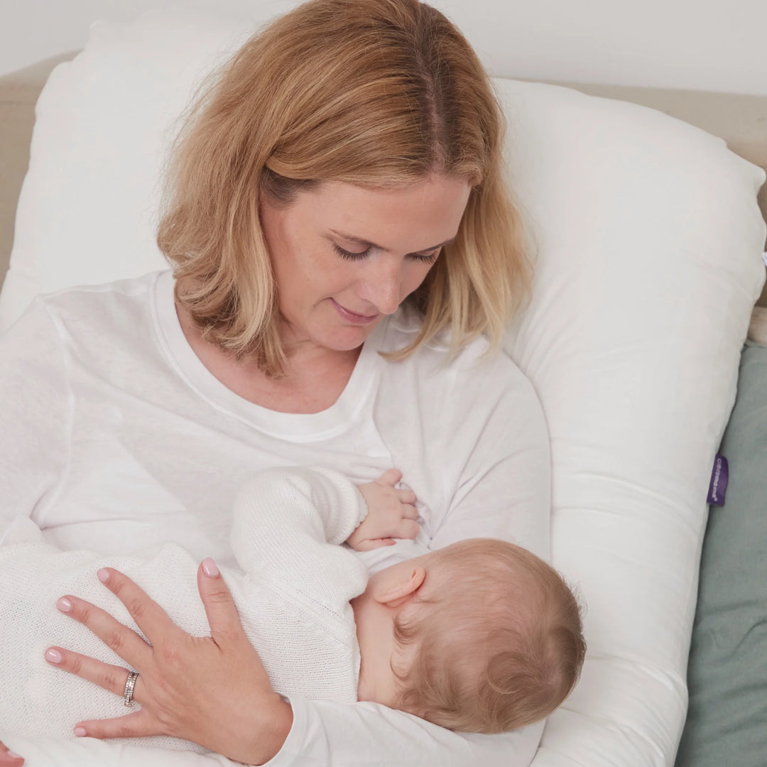 Mother sitting up in bed using a white maternity pillow while breastfeeding her baby, holding the infant close and looking down lovingly