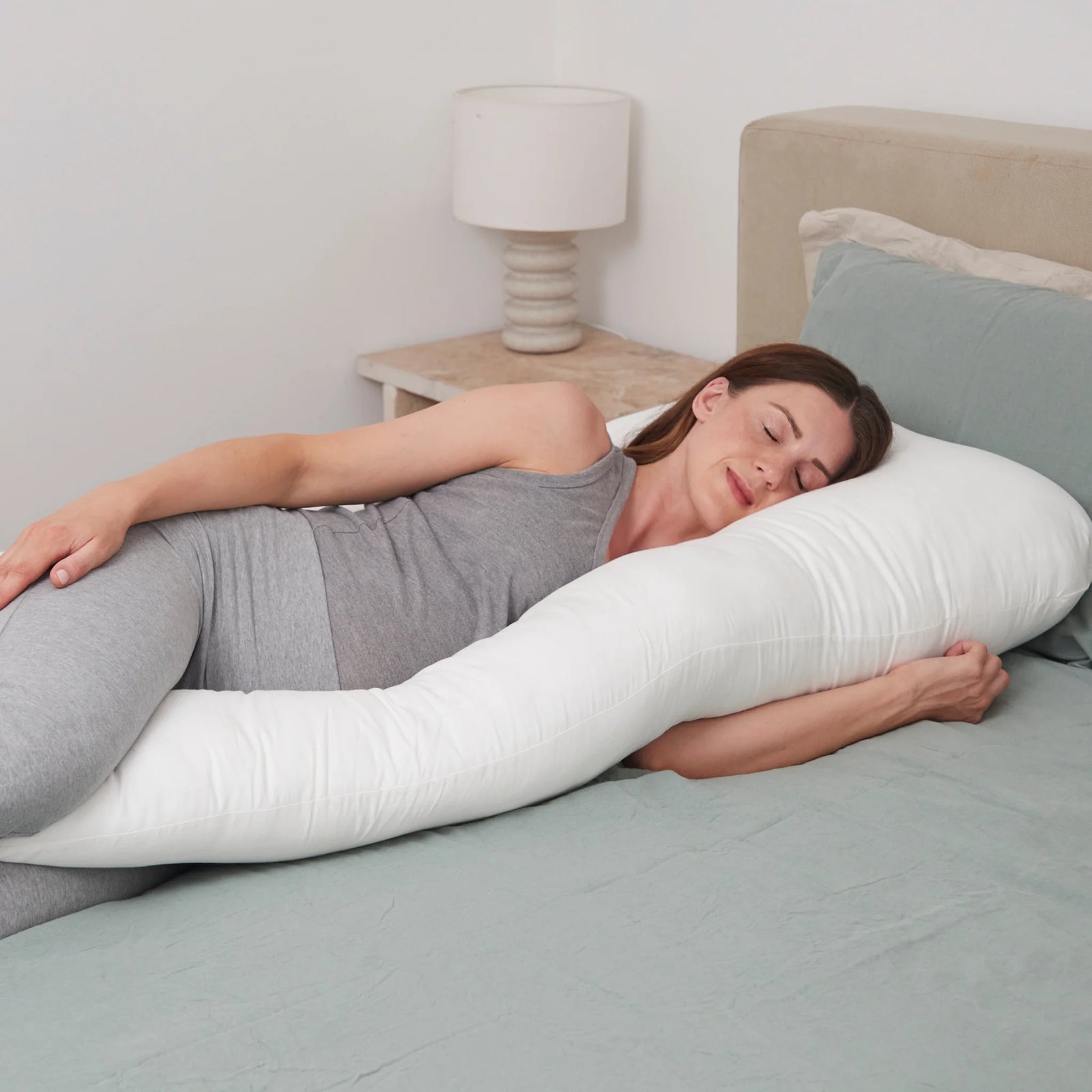 Pregnant woman sleeping comfortably on her side, supported by a large white maternity pillow that cushions her bump, back, and legs.