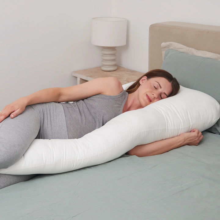 Pregnant woman sleeping comfortably on her side, supported by a large white maternity pillow that cushions her bump, back, and legs.