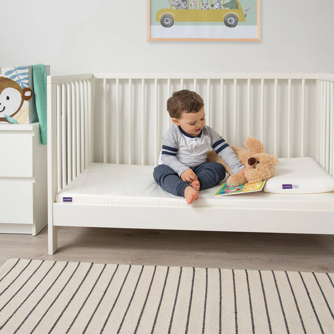 Toddler sitting on Clevamama Baby & Toddler Waterproof Support Mattress in white cot, reading with teddy bear beside him – premium waterproof cot bed mattress with firm reflex foam support, breathable and hypoallergenic cover for safe, comfortable toddler sleep. Ideal transition mattress for baby to toddler bed, asthma and allergy certified.