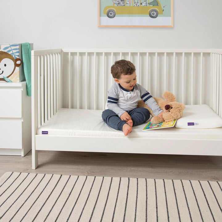 Toddler sitting on Clevamama Baby & Toddler Waterproof Support Mattress in white cot, reading with teddy bear beside him – premium waterproof cot bed mattress with firm reflex foam support, breathable and hypoallergenic cover for safe, comfortable toddler sleep. Ideal transition mattress for baby to toddler bed, asthma and allergy certified.
