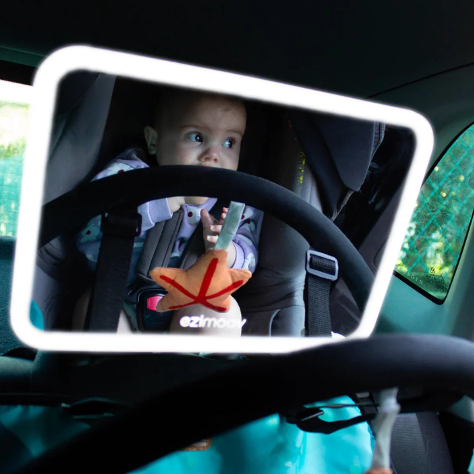 Rear-facing baby in a car seat seen clearly through the illuminated EziMoov LED Pro car seat mirror, showing bright LED frame and secure baby travel setup for safer driving