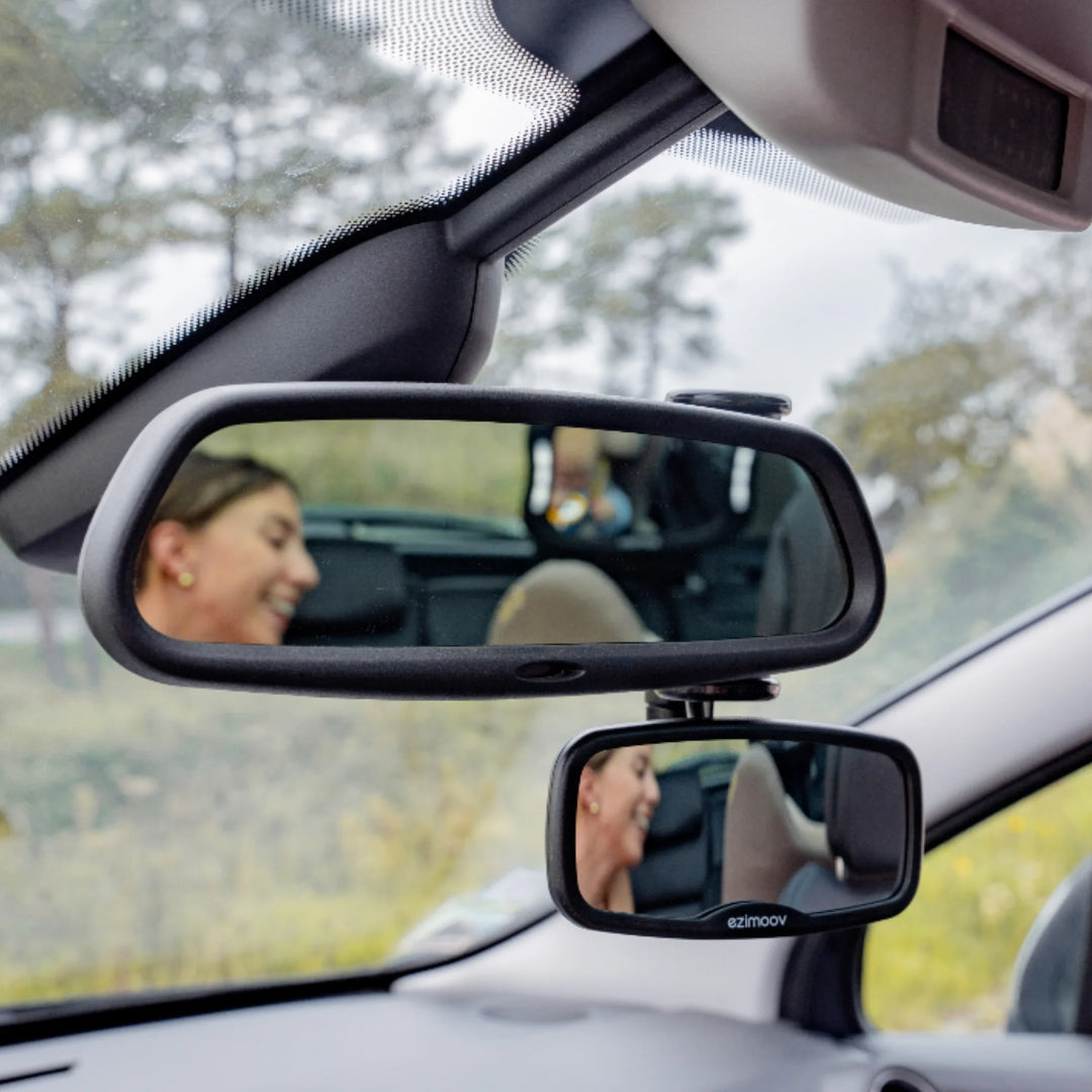Rear-view mirror setup in a car showing an adjustable clip-on baby mirror by Ezimoov, reflecting a smiling parent and child for safe back-seat visibility during driving.