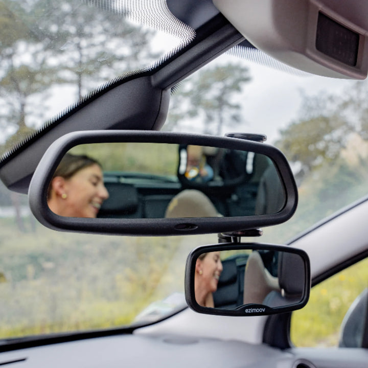 Ezimooov clip-on rear-view baby mirror installed below the main car mirror, reflecting a parent interacting with a child in the back seat; demonstrates easy attachment and clear visibility for monitoring kids while driving.