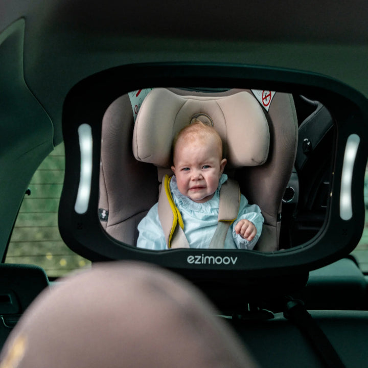 Baby visible in an EziMoov LED rear-facing car seat mirror, showing a clear wide-angle reflection of a crying infant in a beige car seat. LED-illuminated baby car mirror for safer monitoring while driving.