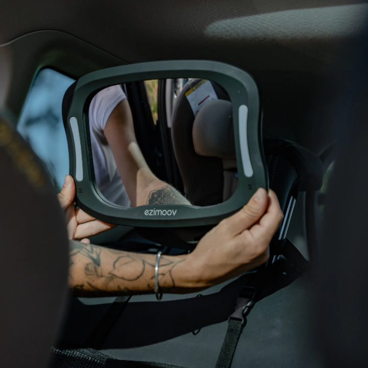 Adult adjusting the EziMoov LED rear-facing baby car seat mirror on a backseat headrest, showing the wide viewing angle and built-in LED lights designed for safer infant monitoring while driving.