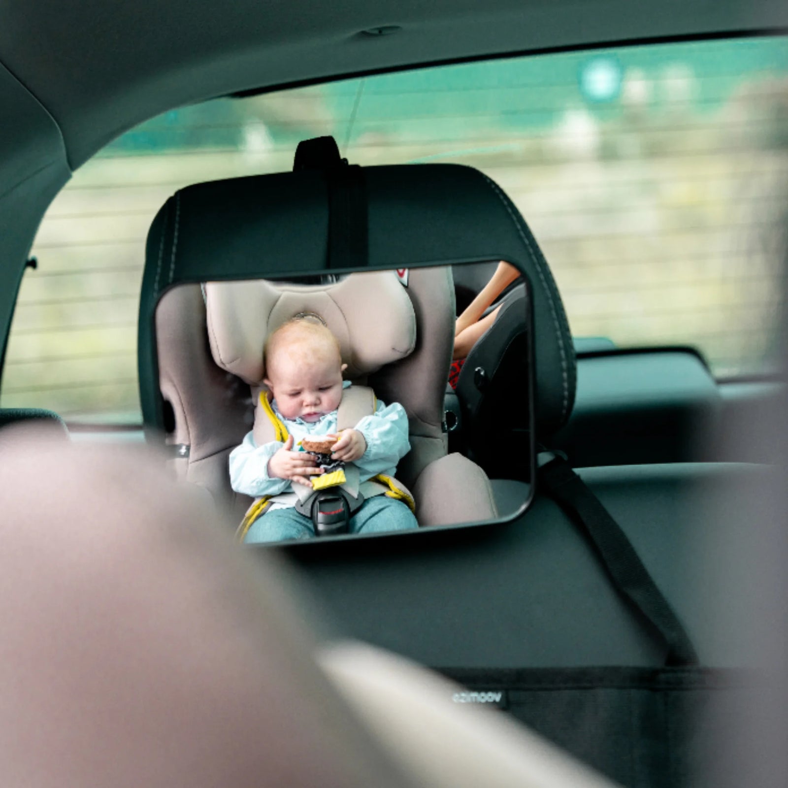 Rear-facing baby car seat mirror showing a clear view of a baby sitting safely in a beige car seat with yellow straps in the back seat of a car. Large wide-angle EziMoov rear-facing car seat mirror for monitoring infants while driving.