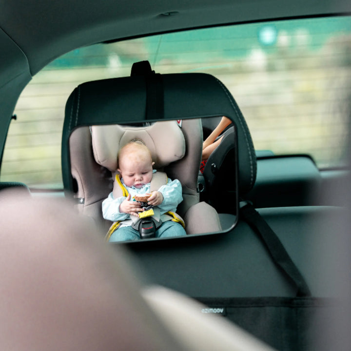 Rear-facing baby car seat mirror showing a clear view of a baby sitting safely in a beige car seat with yellow straps in the back seat of a car. Large wide-angle EziMoov rear-facing car seat mirror for monitoring infants while driving.