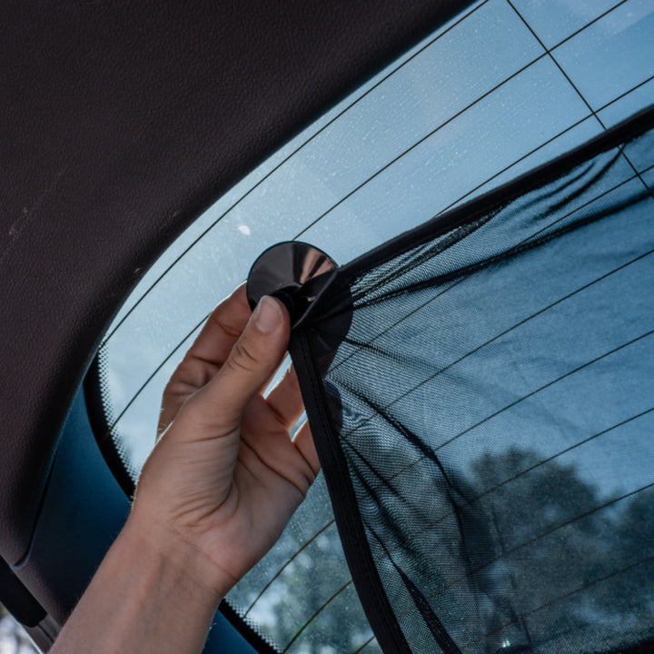 Hand pressing suction cup to attach the EziMoov rear windscreen sunshade to the back window, demonstrating quick and secure installation of the UV-blocking mesh sun shield for car rear windows.