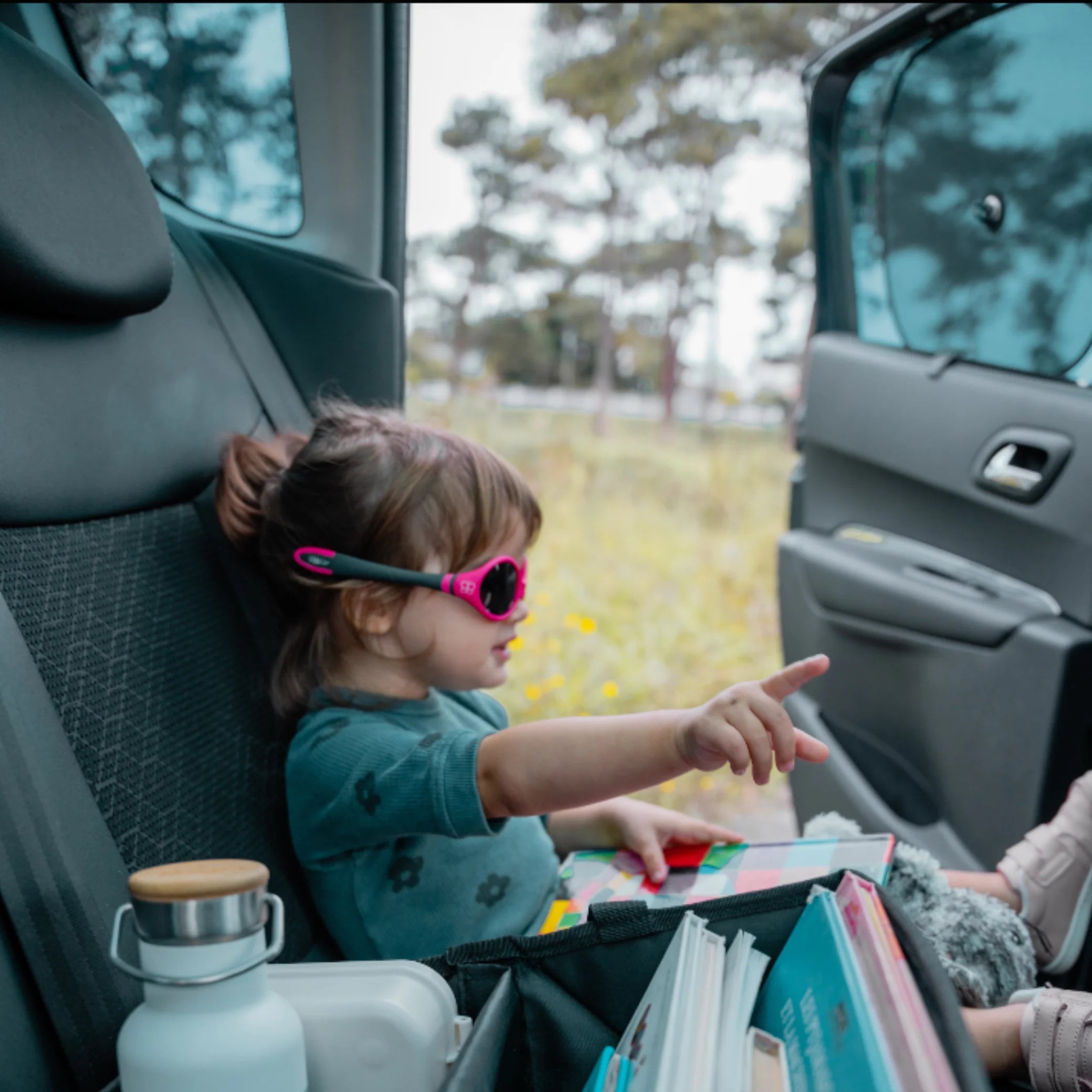 Toddler sitting in the back seat of a car wearing pink sunglasses, reading and pointing while protected from sunlight by an EziMoov car sun shade on the window. Family travel lifestyle image showcasing child comfort and UV protection.