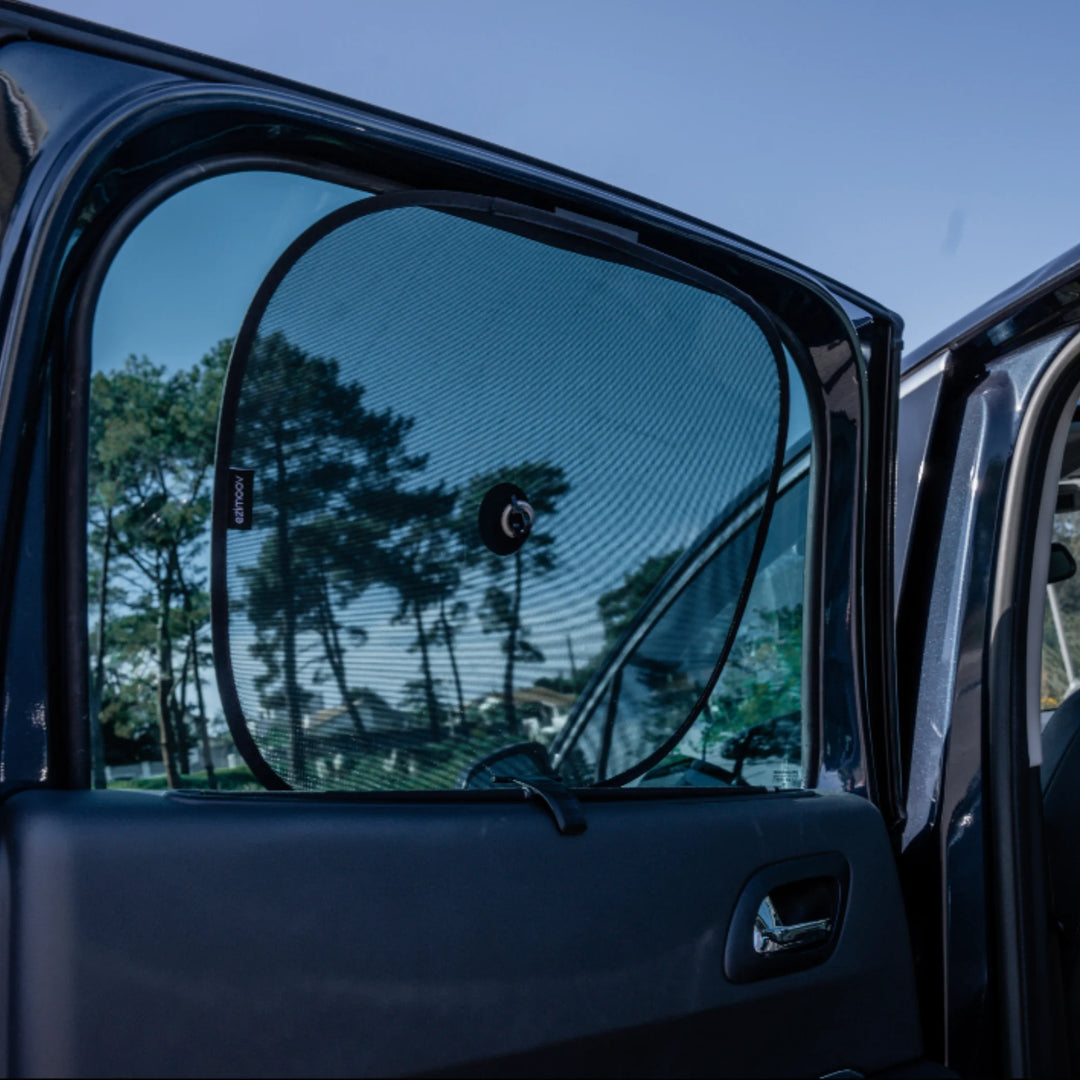 EziMoov car sun shade with central suction cup attached to the rear passenger window, blocking harsh sunlight and providing UV protection for babies and children during car journeys. Outdoor lifestyle shot with car door open.