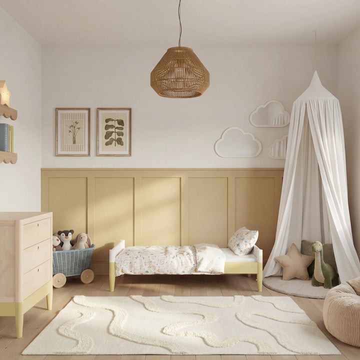 Gaia Baby Eos toddler bed in natural wood and primrose yellow in a warm neutral nursery with canopy, dresser and soft decor.