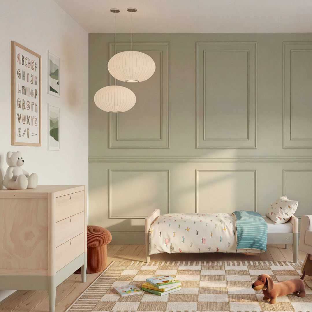 Children’s bedroom with sage green panelled walls, natural wood dresser and toddler bed with colourful bedding in a soft, minimalist nursery design.