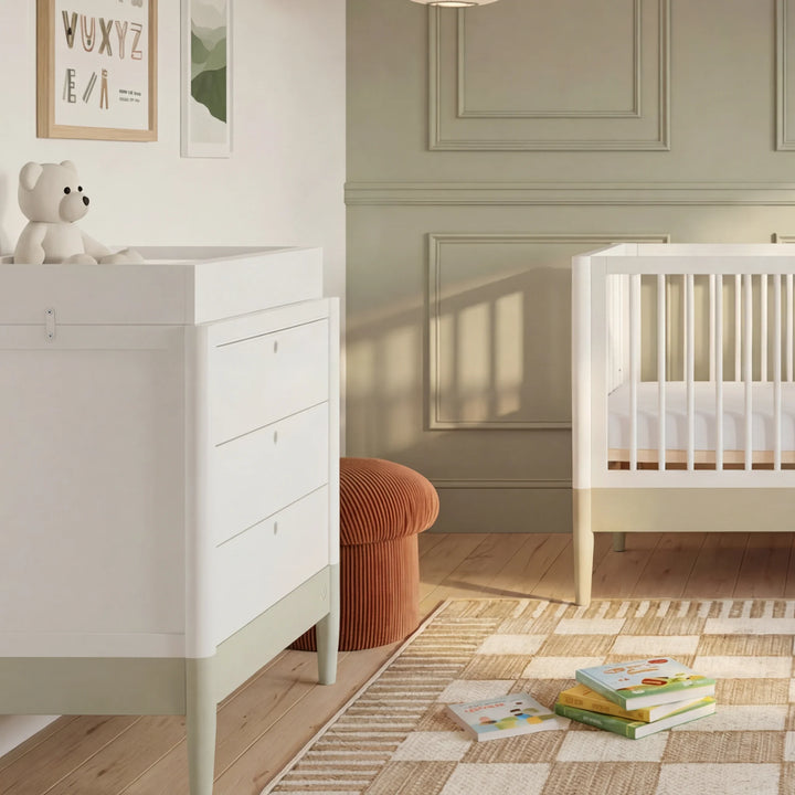 Gaia Baby Eos dresser with changer in white and desert sage in a nursery with cot and books on rug