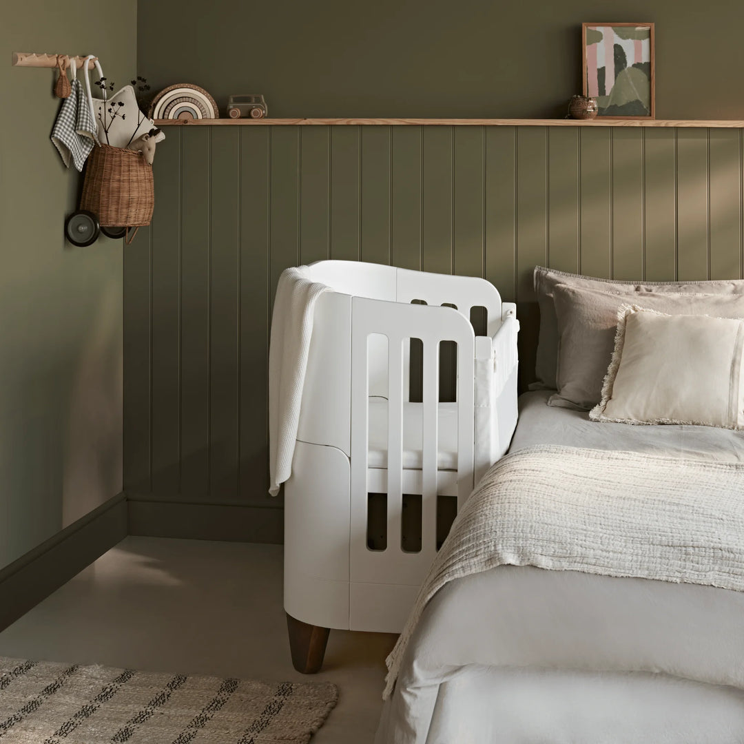 Gaia Baby Serena bedside crib add-on in white with walnut legs attached to a bed, side view in a nursery with green paneled walls