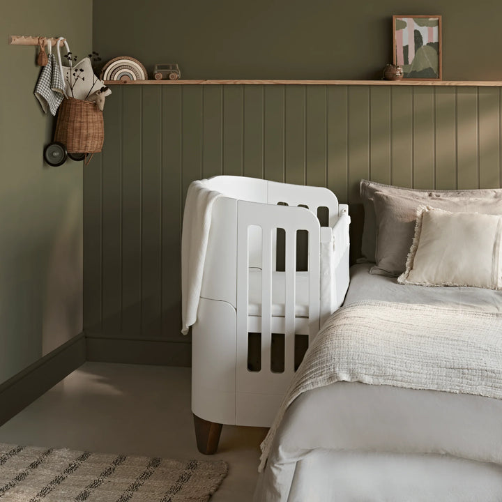Gaia Baby Serena bedside crib add-on in white with walnut legs attached to a bed, side view in a nursery with green paneled walls