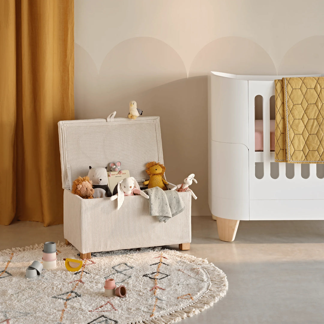 An ecru corduroy storage footstool styled as a toy chest in a warm nursery setting, filled with soft teddy bears and plush toys beside a modern white crib, mustard curtains, and a textured rug for a cozy and playful atmosphere.