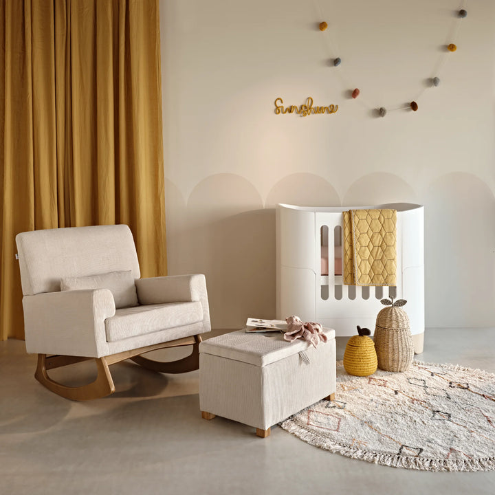 A cozy nursery corner featuring the Gaia Baby Serena rocking chair and matching footstool in ecru corduroy fabric beside a white crib, with warm mustard curtains, soft neutral décor, woven baskets, and a textured rug creating a calm and stylish nursery setting.