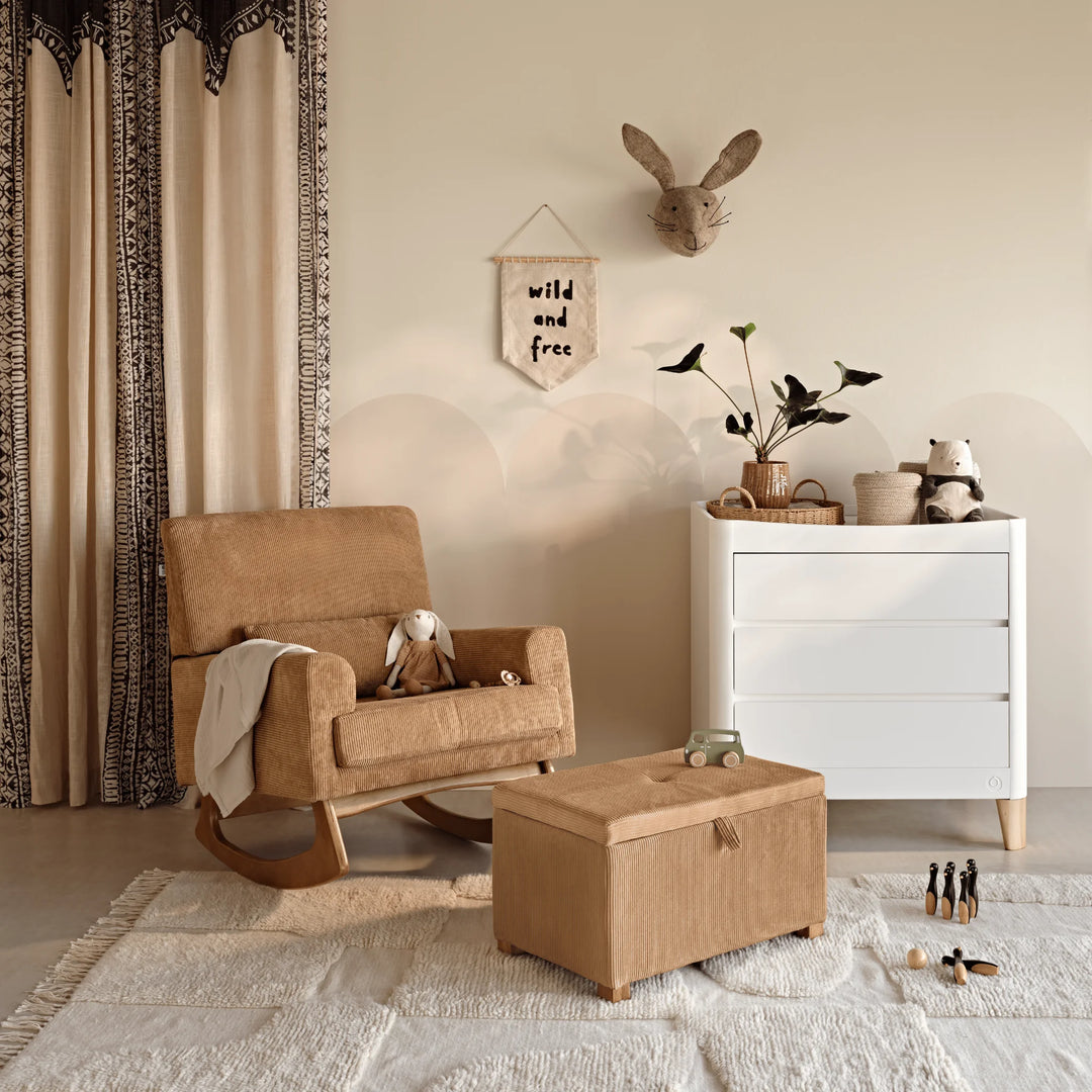 A warm, neutral-toned nursery featuring the Gaia Baby Serena rocking nursing chair and matching storage footstool in caramel corduroy fabric, paired with a white dresser, soft textiles, and playful décor for a cosy contemporary nursery setting.