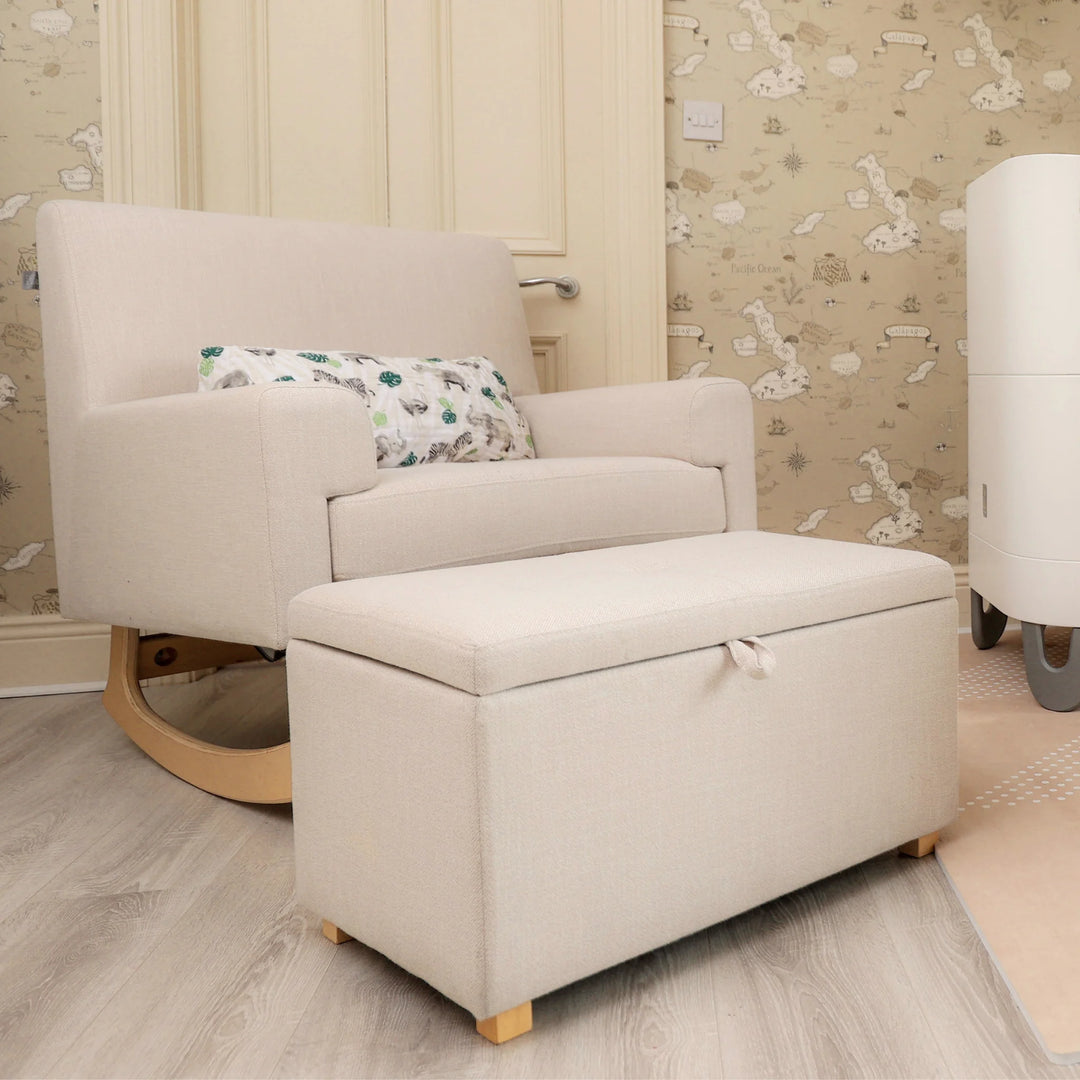 The Gaia Baby Serena rocking chair and matching storage footstool in soft oat fabric, styled together in a cozy nursery with warm wallpaper and natural wood accents, offering comfortable seating and practical hidden storage for modern family spaces.