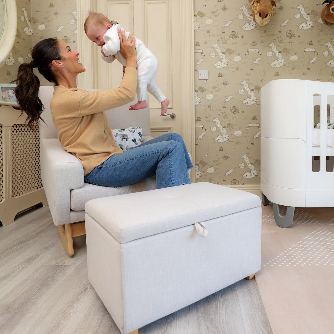 A mother sitting in a cozy nursery on the Gaia Baby Serena nursing chair, lifting her baby with joy beside the matching storage footstool and a modern white crib, creating a warm and comforting family moment.