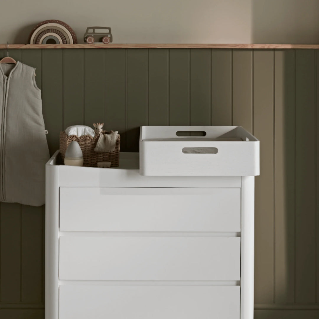 Gaia Baby Hera white changing station displayed on matching dresser in a cosy neutral nursery, styled with woven baskets, baby essentials and warm wooden wall panelling.