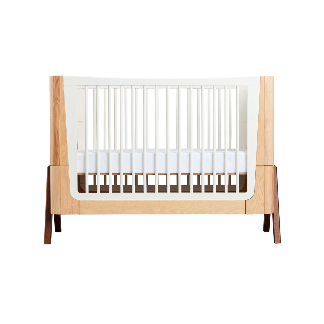 Front view of the Gaia Baby Hera cot in ash and walnut, featuring curved Scandinavian design, natural wood panels, white slatted sides and solid walnut A-frame legs — premium sustainable baby crib.