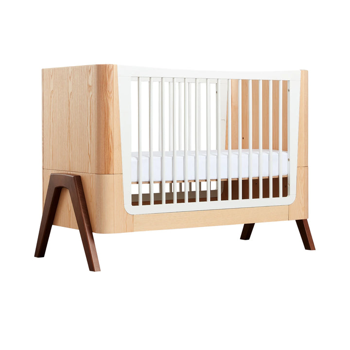 Gaia Baby Hera cot in ash and walnut, angled side view showing curved Scandinavian design, natural wood panels, white slatted sides and solid walnut A-frame legs — premium sustainable baby crib.