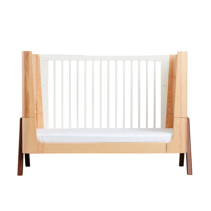 Gaia Baby Hera cot in ash and walnut converted into toddler bed mode, featuring Scandinavian curved design, natural wood panels, white slatted side and solid walnut A-frame legs — premium sustainable nursery furniture.