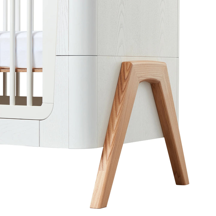 Close-up of Gaia Baby Hera cot bed in Scandi White showing natural solid wood A-frame legs, curved modern Scandinavian detailing and premium sustainable nursery furniture design.