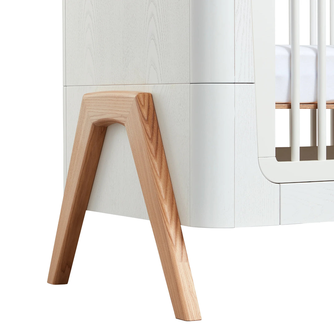 Close-up of Gaia Baby Hera cot bed in Scandi White showing natural solid wood A-frame legs and smooth curved Scandinavian detailing — premium sustainable nursery furniture craftsmanship.