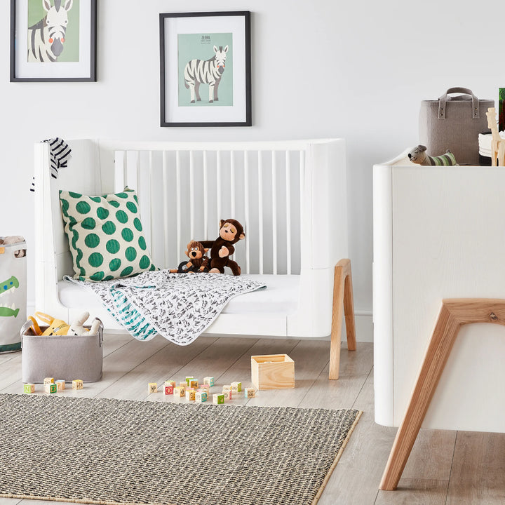 Lifestyle nursery scene featuring the Gaia Baby Hera cot bed in Scandi White with natural wood legs, styled as a toddler daybed with cushions, blankets and toys — modern Scandinavian nursery furniture in a bright contemporary baby room
