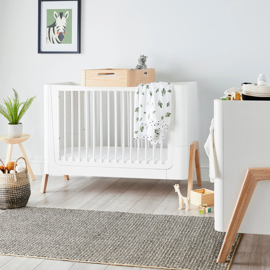 Bright modern nursery featuring the Gaia Baby Hera cot bed in Scandi White with natural wood legs, styled with neutral décor, toys and greenery — Scandinavian-inspired sustainable baby furniture in a contemporary room setting.