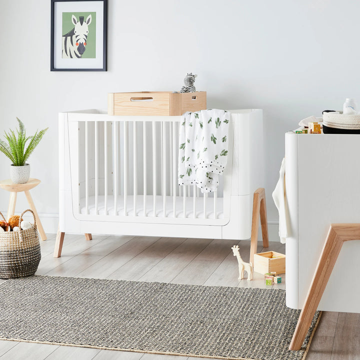 Bright modern nursery featuring the Gaia Baby Hera cot bed in Scandi White with natural wood legs, styled with neutral décor, toys and greenery — Scandinavian-inspired sustainable baby furniture in a contemporary room setting.