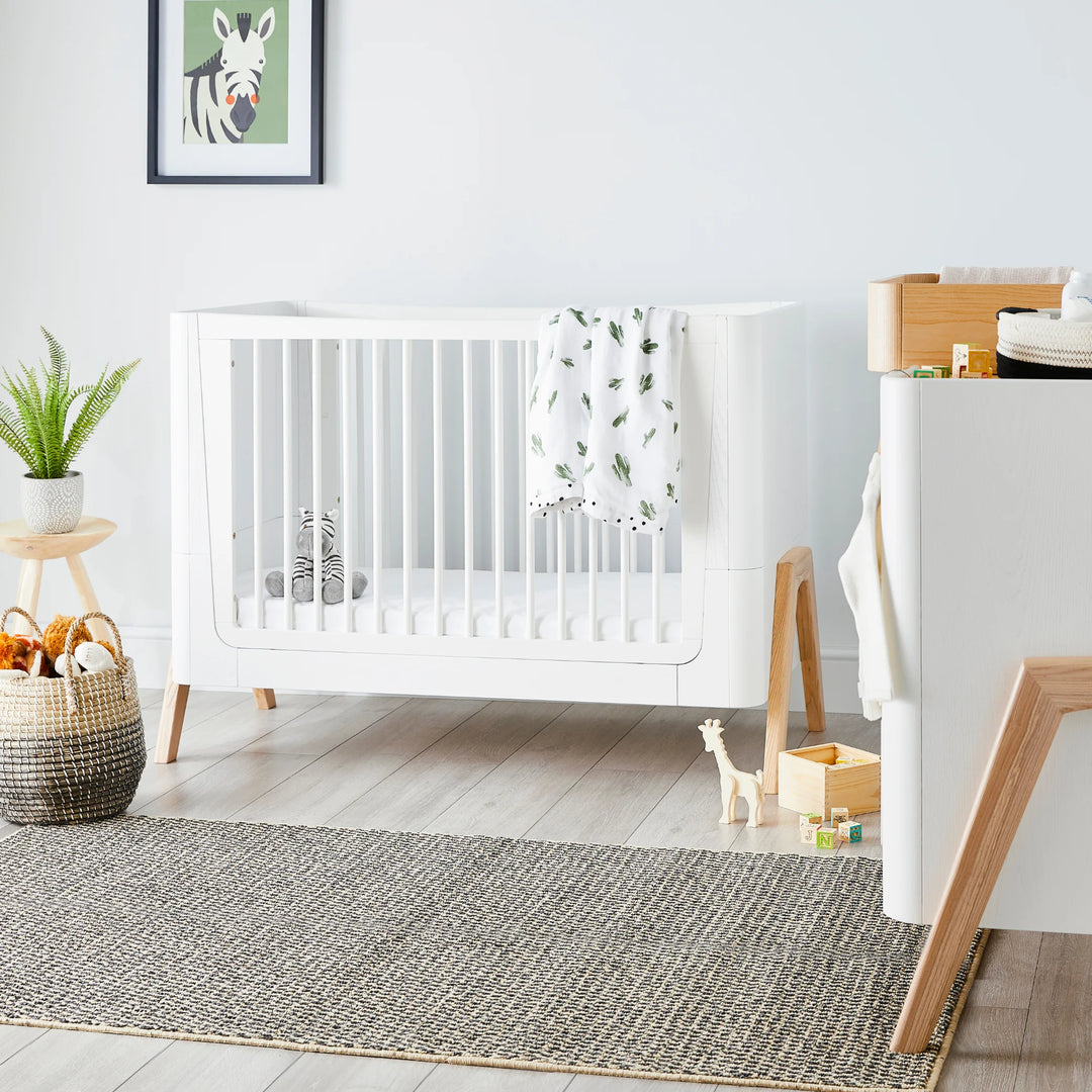 Modern nursery room featuring the Gaia Baby Hera cot bed in Scandi White with natural wood legs, styled with soft neutral décor, houseplants, toys and a woven storage basket — premium Scandinavian sustainable baby furniture.