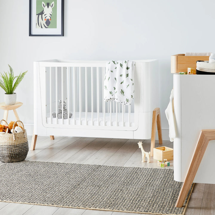 Modern nursery room featuring the Gaia Baby Hera cot bed in Scandi White with natural wood legs, styled with soft neutral décor, houseplants, toys and a woven storage basket — premium Scandinavian sustainable baby furniture.