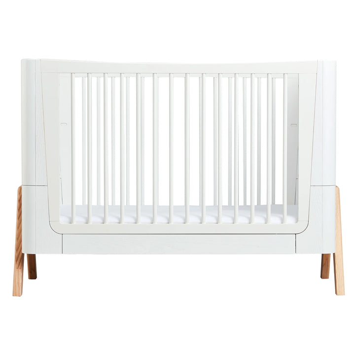 Gaia Baby Hera cot bed in Scandi White with natural wood legs, shown with mattress at lowest height, featuring curved Scandinavian design and white slatted sides — premium sustainable convertible baby cot.